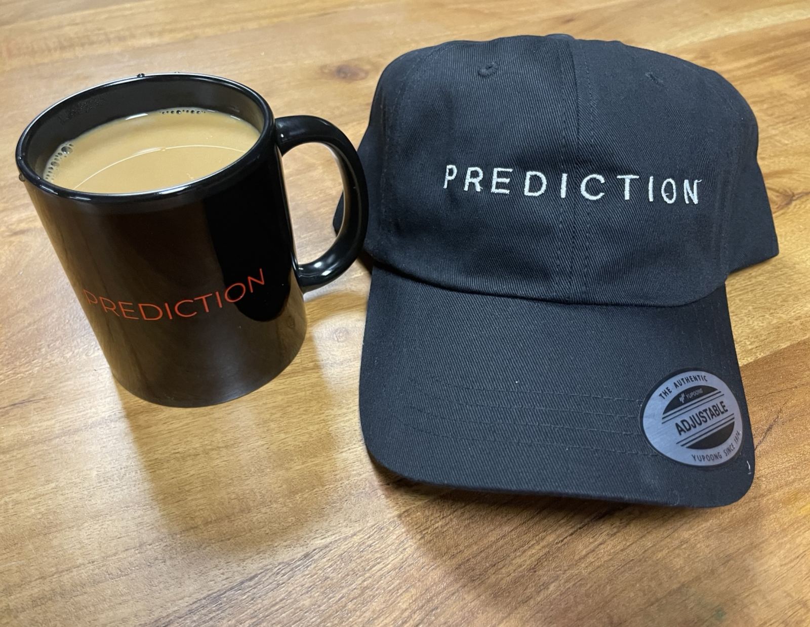 A black Prediction coffee mug and black Prediction cap on a wooden table